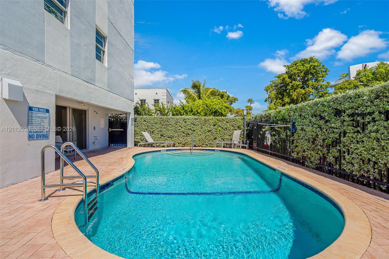 In-ground swimming pool featuring a curved design, metal handrail, concrete paver decking, and surrounding privacy hedging