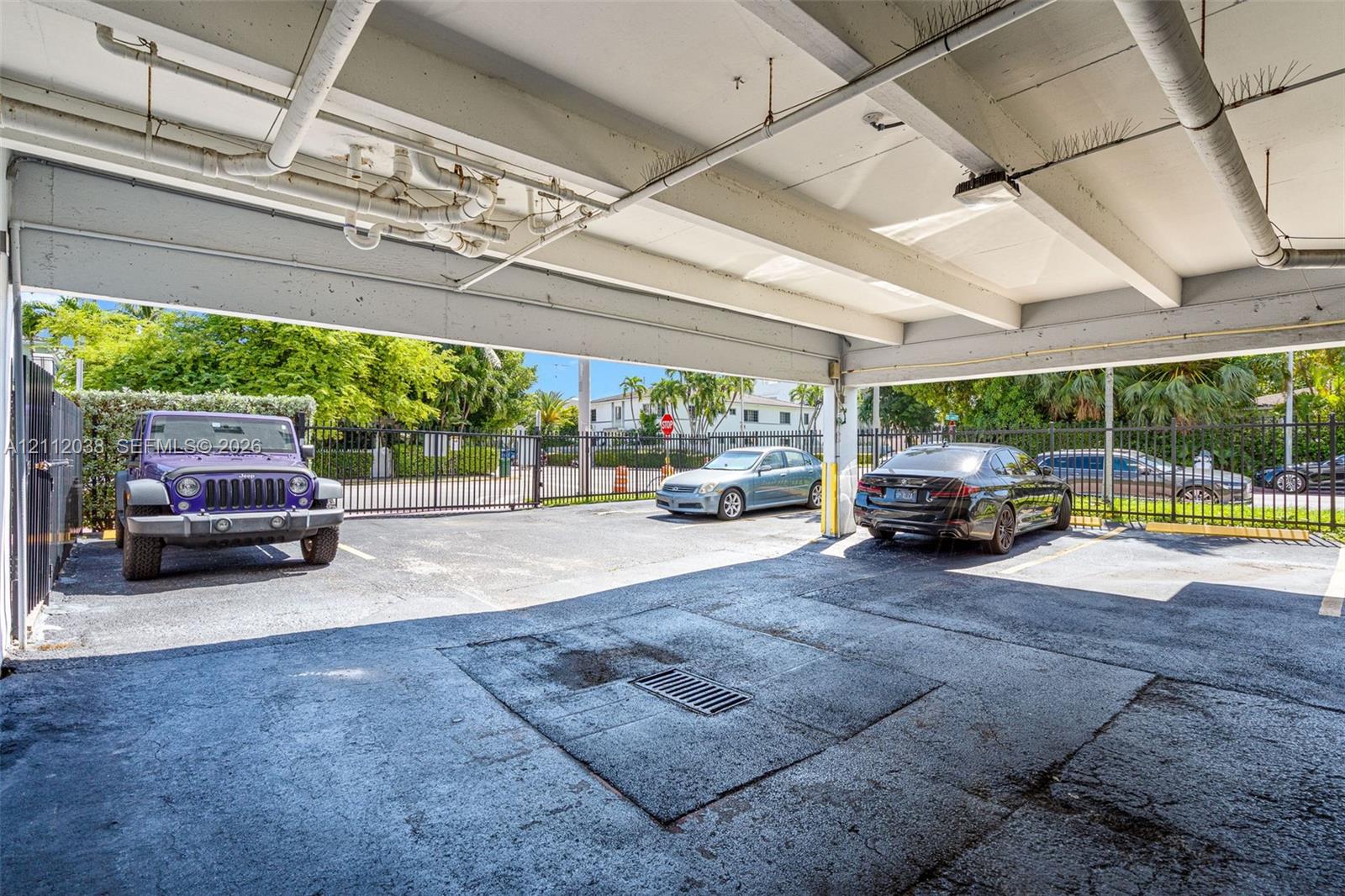 Covered parking area featuring a concrete ceiling structure, asphalt flooring, drainage grate, and perimeter metal fencing