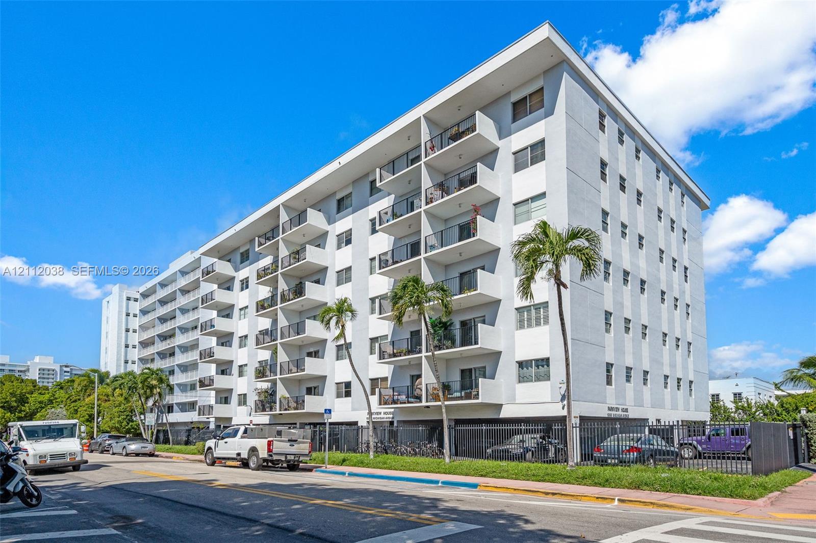 Mid-rise residential building featuring a concrete facade, tiered private balconies with metal railings, and secured ground-level parking