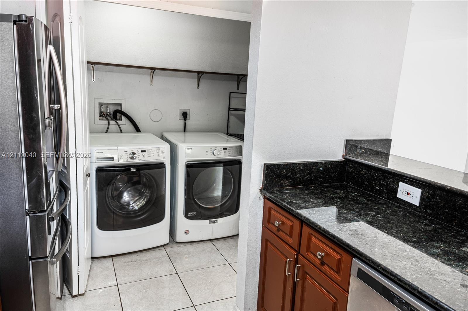 Laundry area with built-in shelving, dark stone-finish countertops, warm-toned wood cabinetry, and tiled flooring