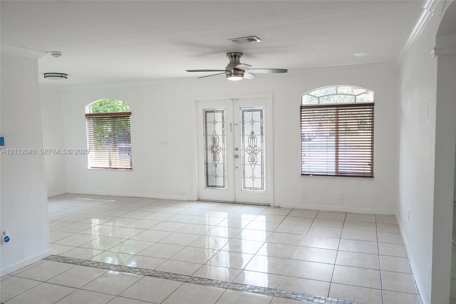 Open-concept interior featuring light-toned tile flooring, double entry doors with decorative glass inserts, arched windows, and a ceiling fan with integrated lighting