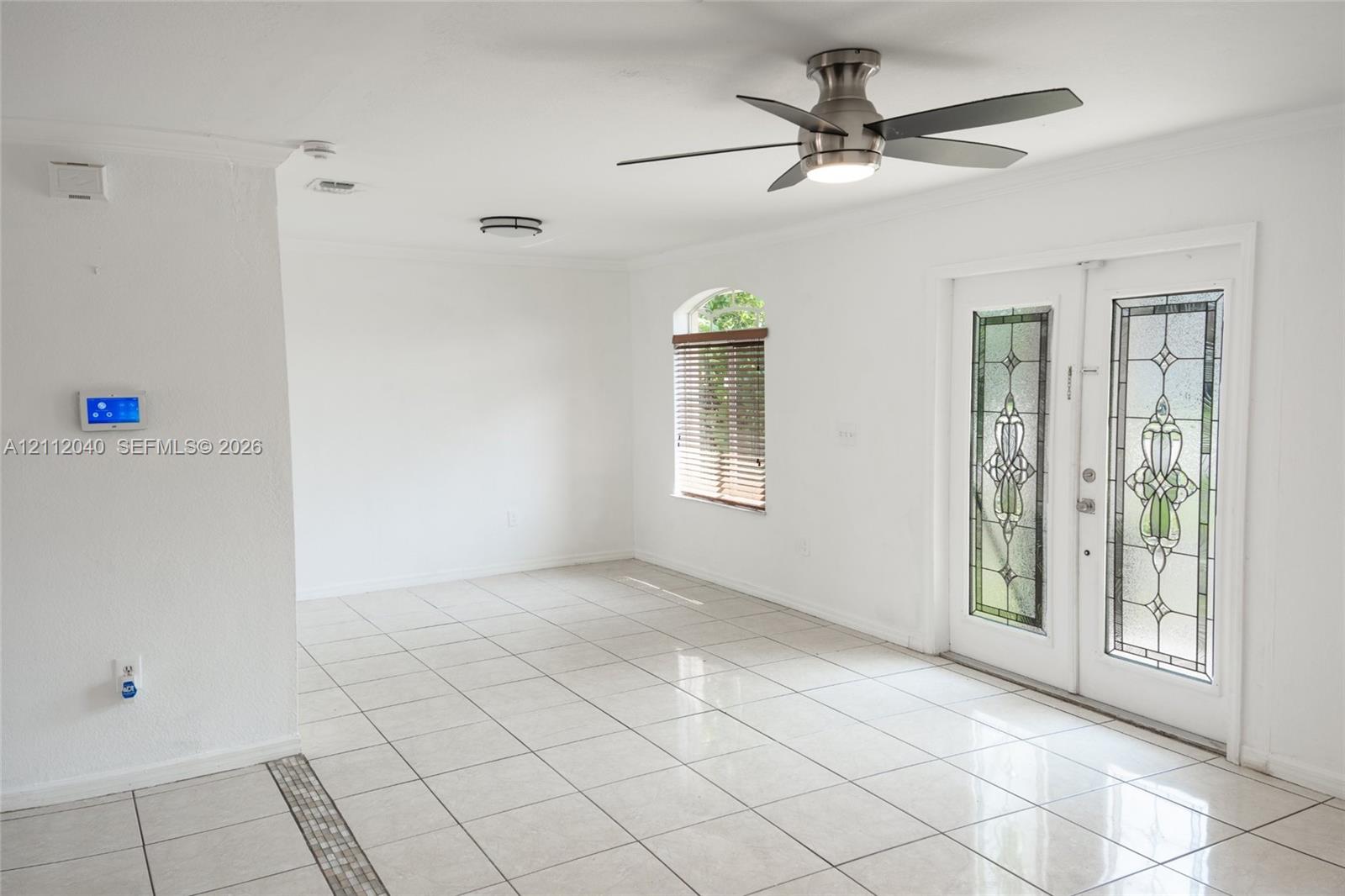 Light-filled room featuring large-format tile flooring, dual entry doors with decorative glass inserts, an arched window with horizontal blinds, and a modern ceiling fan with integrated lighting