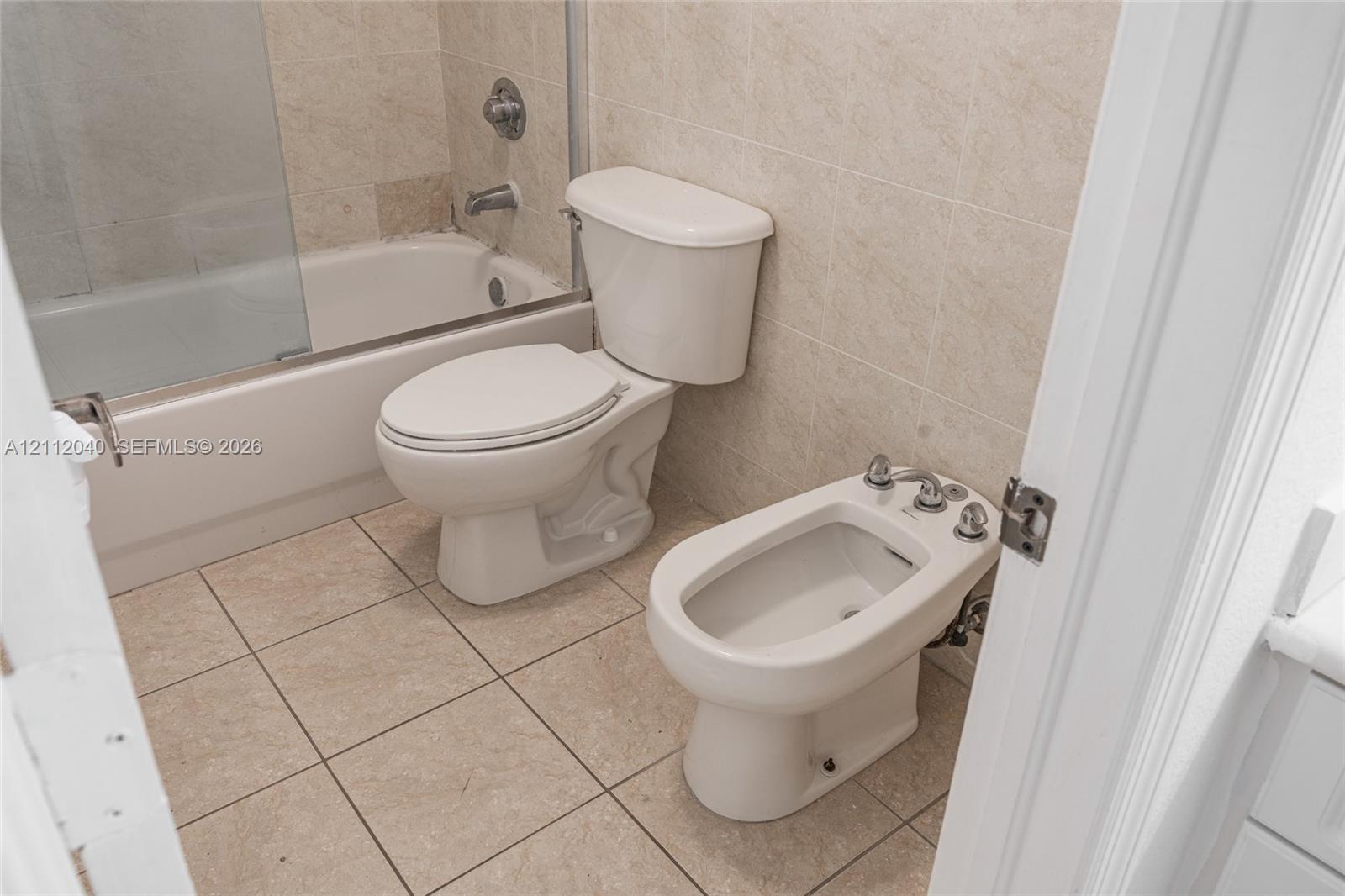 Bathroom featuring a built-in bidet, a two-piece toilet, a porcelain-surround tub with a glass screen, and neutral stone-finish ceramic tile flooring and wall accents