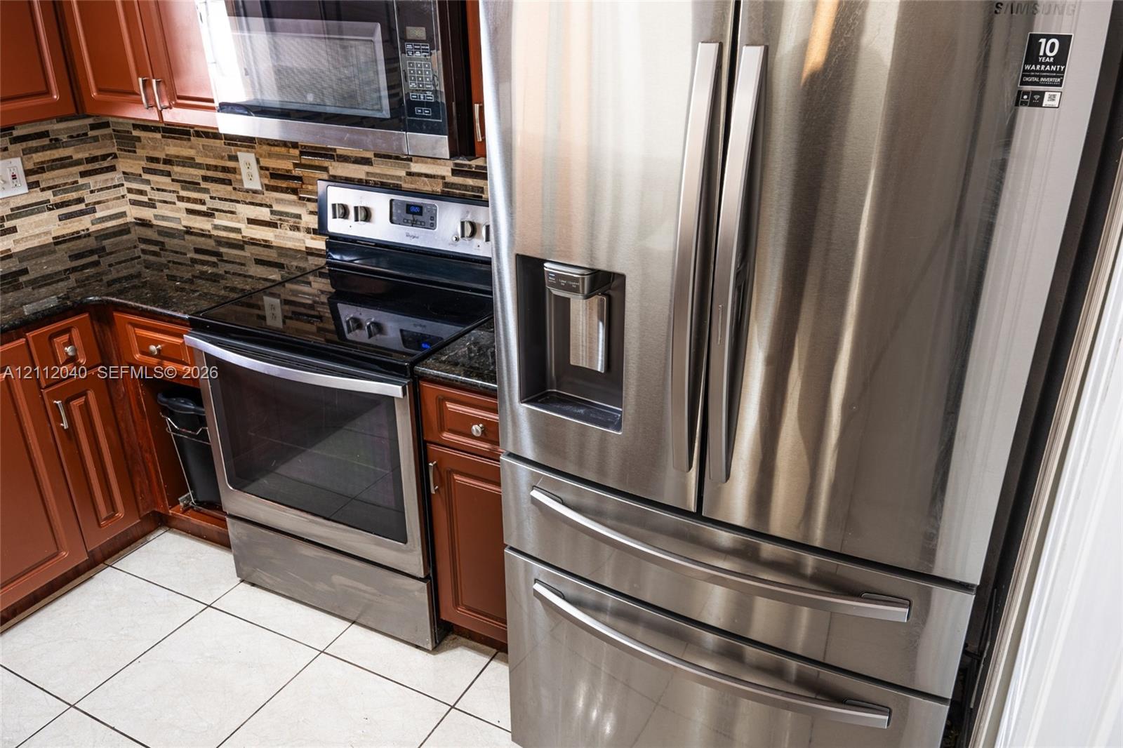 Kitchen featuring stainless steel appliances, dark stone countertops, natural wood-finish cabinetry, mosaic tile backsplash, and ceramic tile flooring