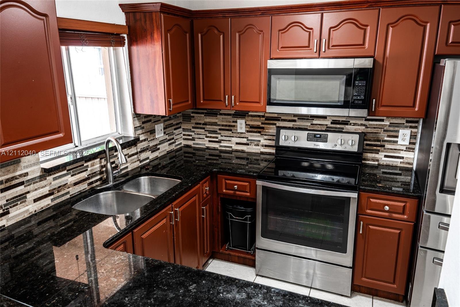 Kitchen featuring warm-toned wood cabinetry, dark stone countertops, stainless steel appliances, and a multi-tone mosaic tile backsplash