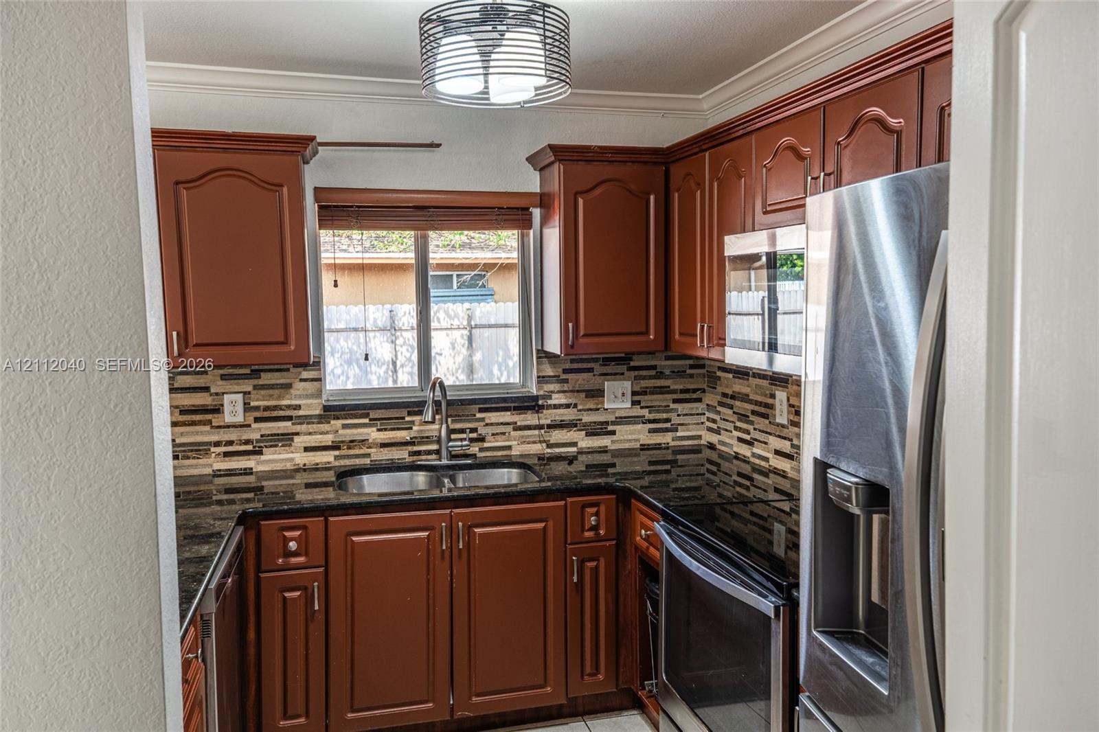 Kitchen featuring stained wood cabinetry, stone countertops, multi-toned tile backsplash, stainless steel appliances, and a circular drum pendant light fixture
