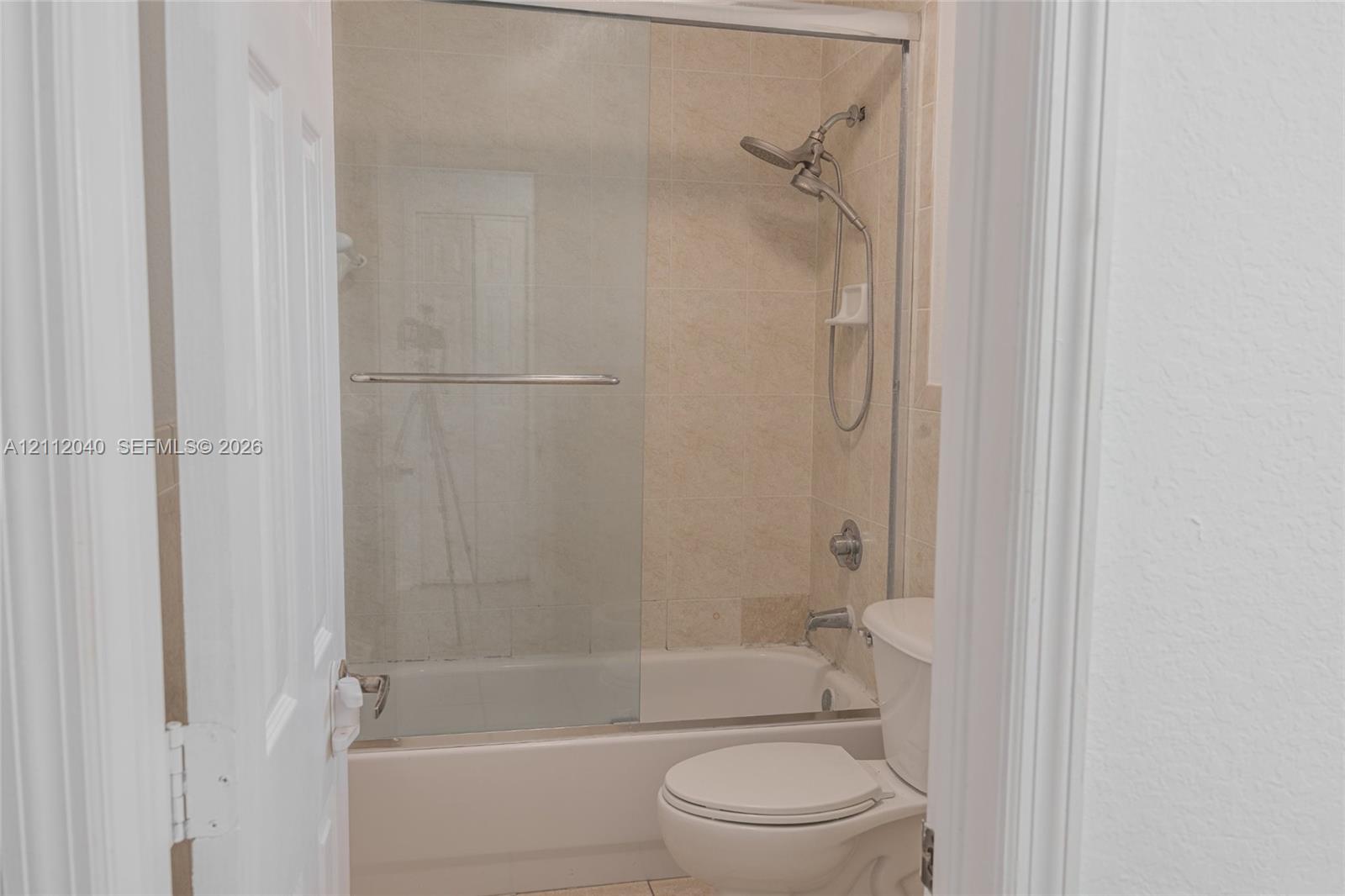Bathroom featuring a tub-shower combination with sliding glass doors, neutral tile surround, adjustable handheld shower fixture, and a white toilet