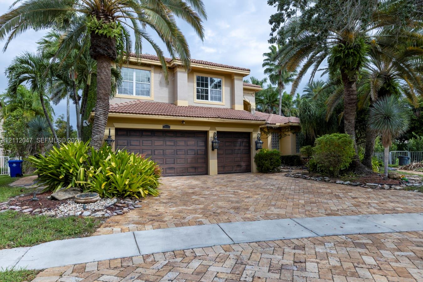 Two-story exterior featuring a tile roof, dual-bay garage, herringbone-patterned stone paver driveway, and mature landscaping
