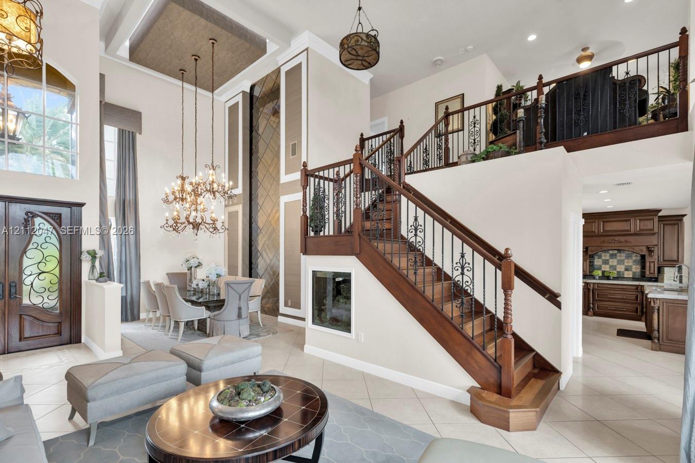 Grand foyer featuring double-height ceilings, a wood staircase with ornate metal balusters, a coffered ceiling with inset paneling, decorative arched glass entry door, and large-scale chandelier lighting
