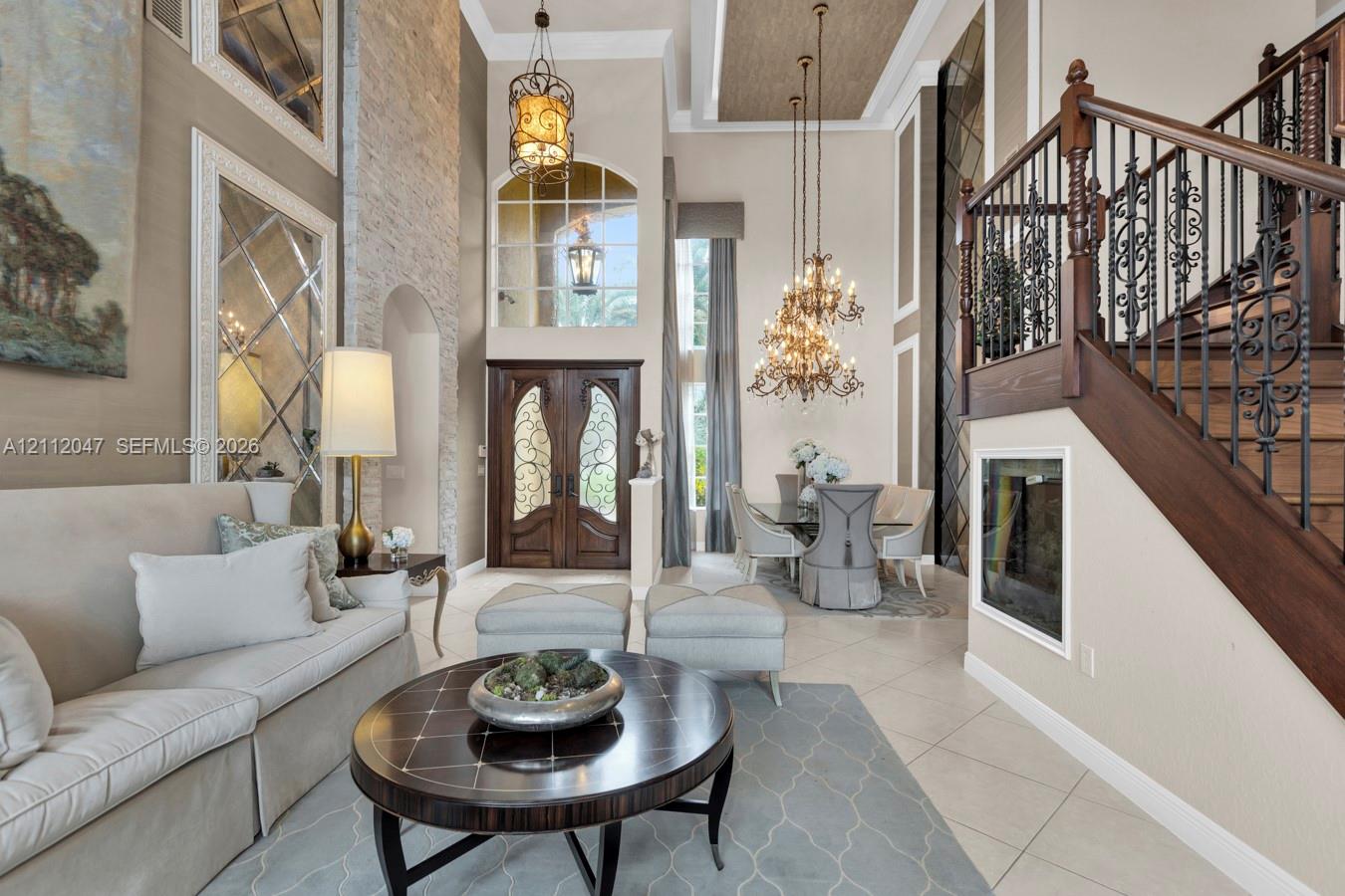 Double-height foyer featuring a stacked stone accent wall, decorative arched wood-paneled front doors with wrought iron detailing, large-scale custom chandeliers, and a grand staircase with dark wood handrails and ornate metal balusters