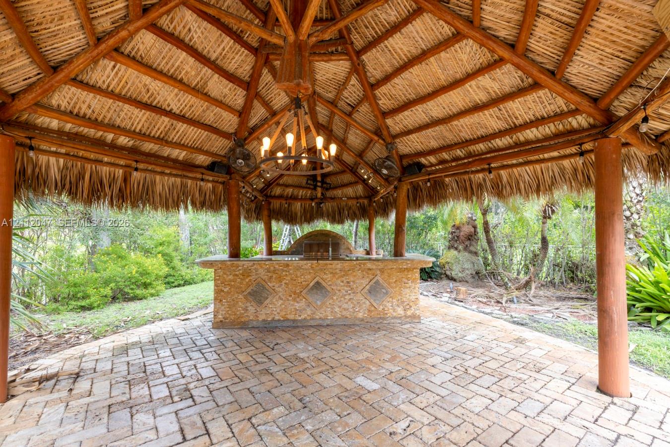 Outdoor palapa featuring a natural thatch roof, exposed wood structural supports, brick paver flooring, built-in stone-finish bar, and a hanging wagon-wheel style fixture
