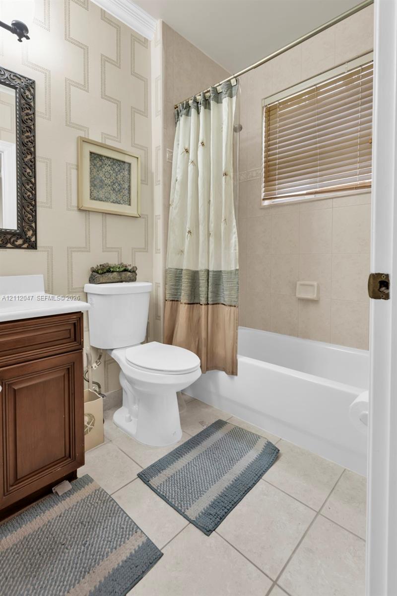 Bathroom featuring a wood-finish vanity with a white countertop, patterned wall covering, tile flooring, and a tub-shower combination with full-height tile surround