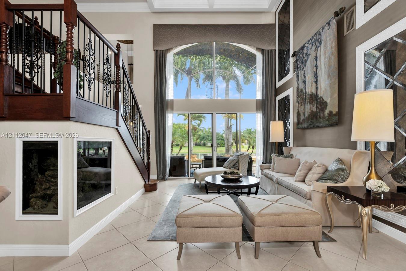 Double-height vaulted ceilings featuring floor-to-ceiling windows, tile flooring, and a decorative staircase with dark wood handrails and wrought iron balusters