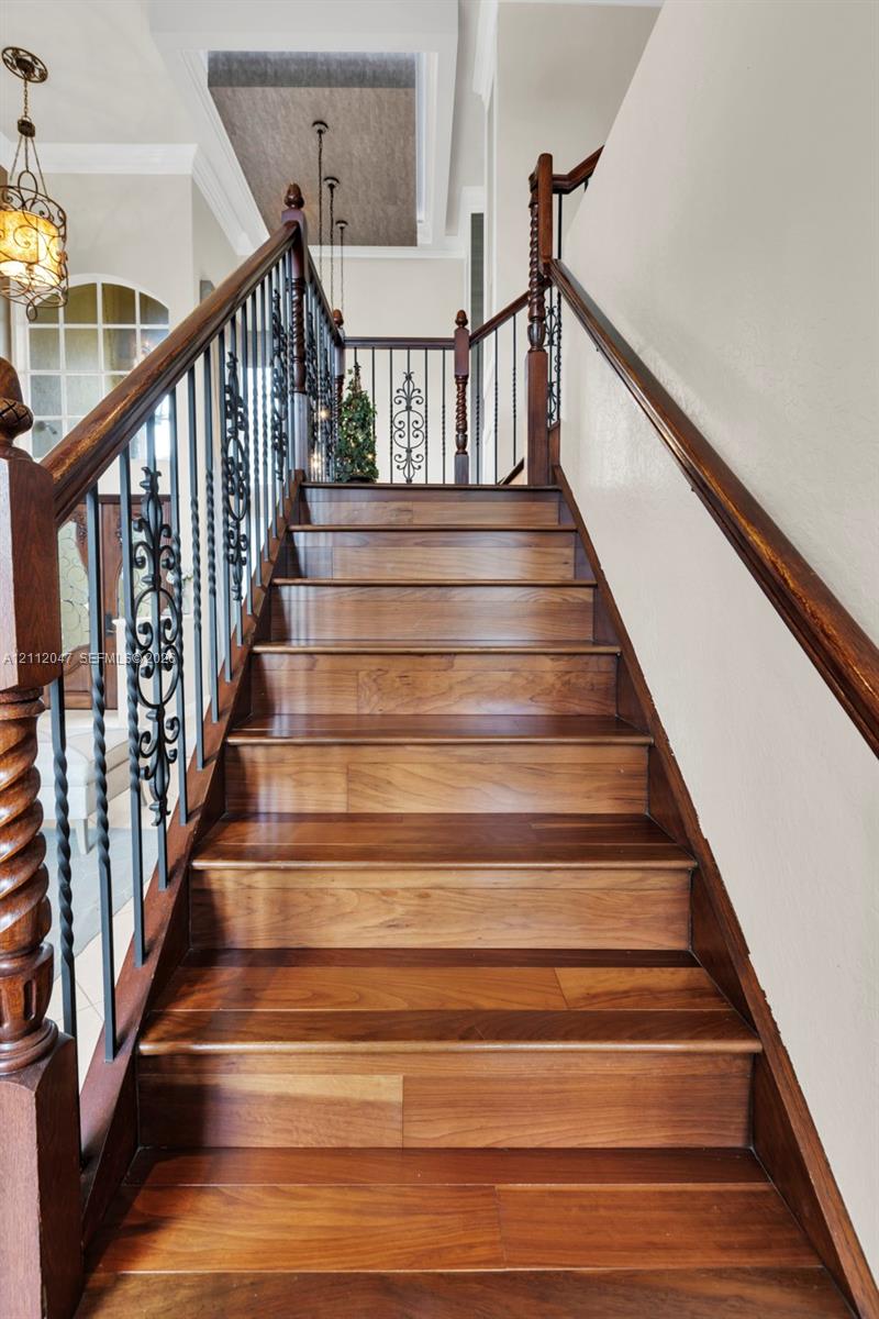 Staircase featuring wood-finish treads, decorative metal balusters, wooden handrails with carved newel posts, and a recessed ceiling detail with pendant lighting