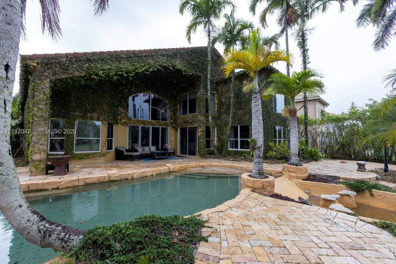Exterior featuring vine-covered masonry, swimming pool with natural stone coping, expansive paver patio, and integrated tropical landscaping