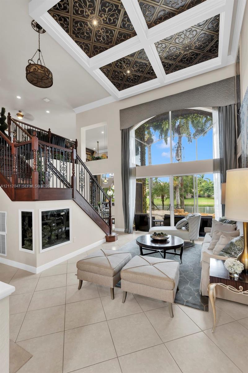 Coffered ceiling with decorative inset panels, floor-to-ceiling windows, ceramic tile flooring, and a mahogany-toned staircase with ornate metal balusters