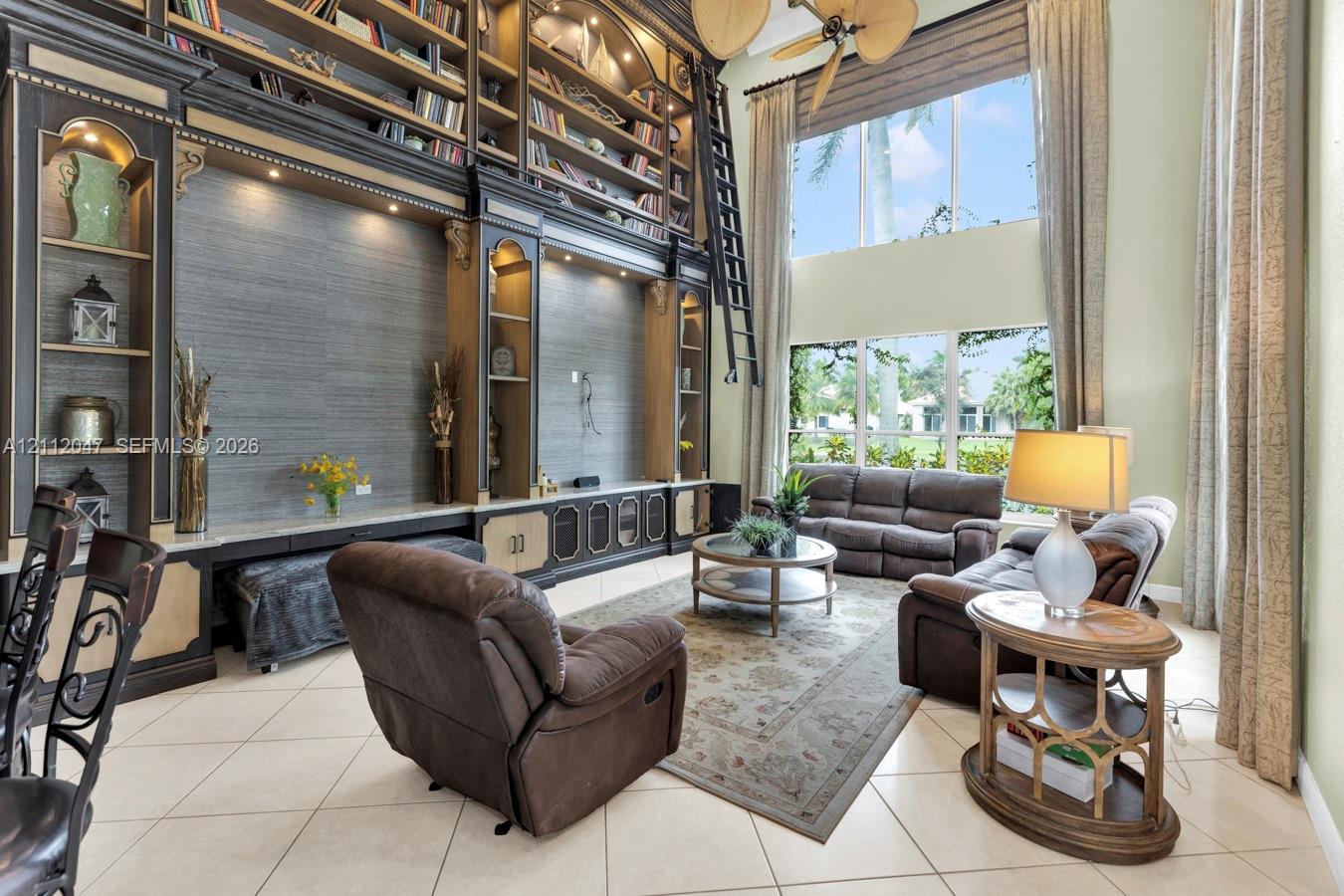 Double-height living area featuring floor-to-ceiling custom built-in cabinetry, integrated library shelving with rolling ladder, oversized windows, and neutral tile flooring