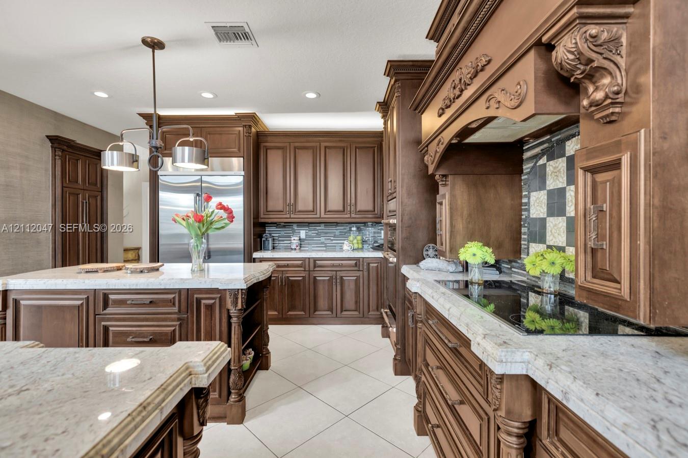 Kitchen featuring custom wood cabinetry with ornate corbels, stone countertops, a large center island, tile flooring, and a decorative tile backsplash