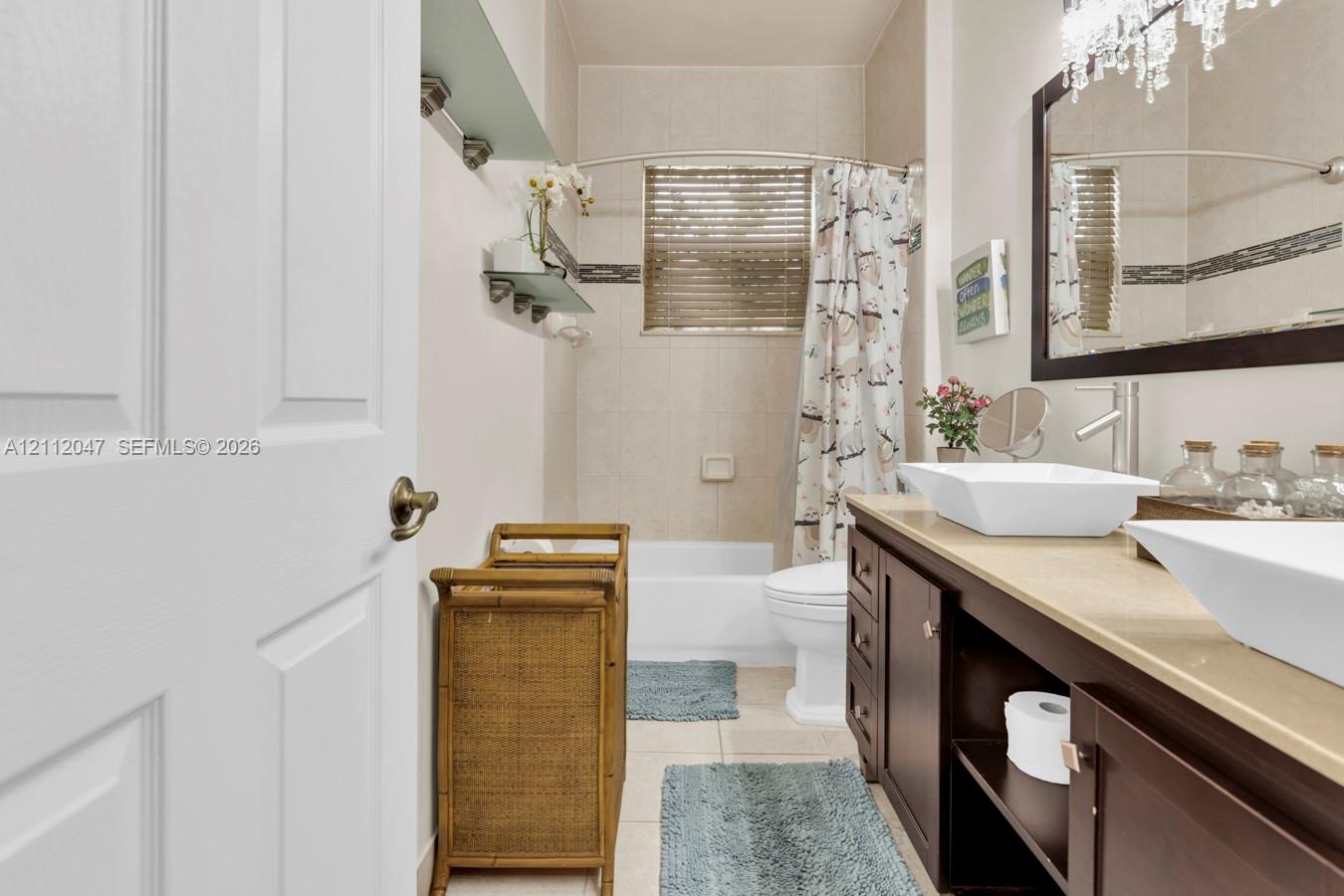 Dark wood vanity featuring dual rectangular vessel sinks, stone-finish countertop, decorative glass wall shelving, and a tiled shower-tub combination with a glass accent border
