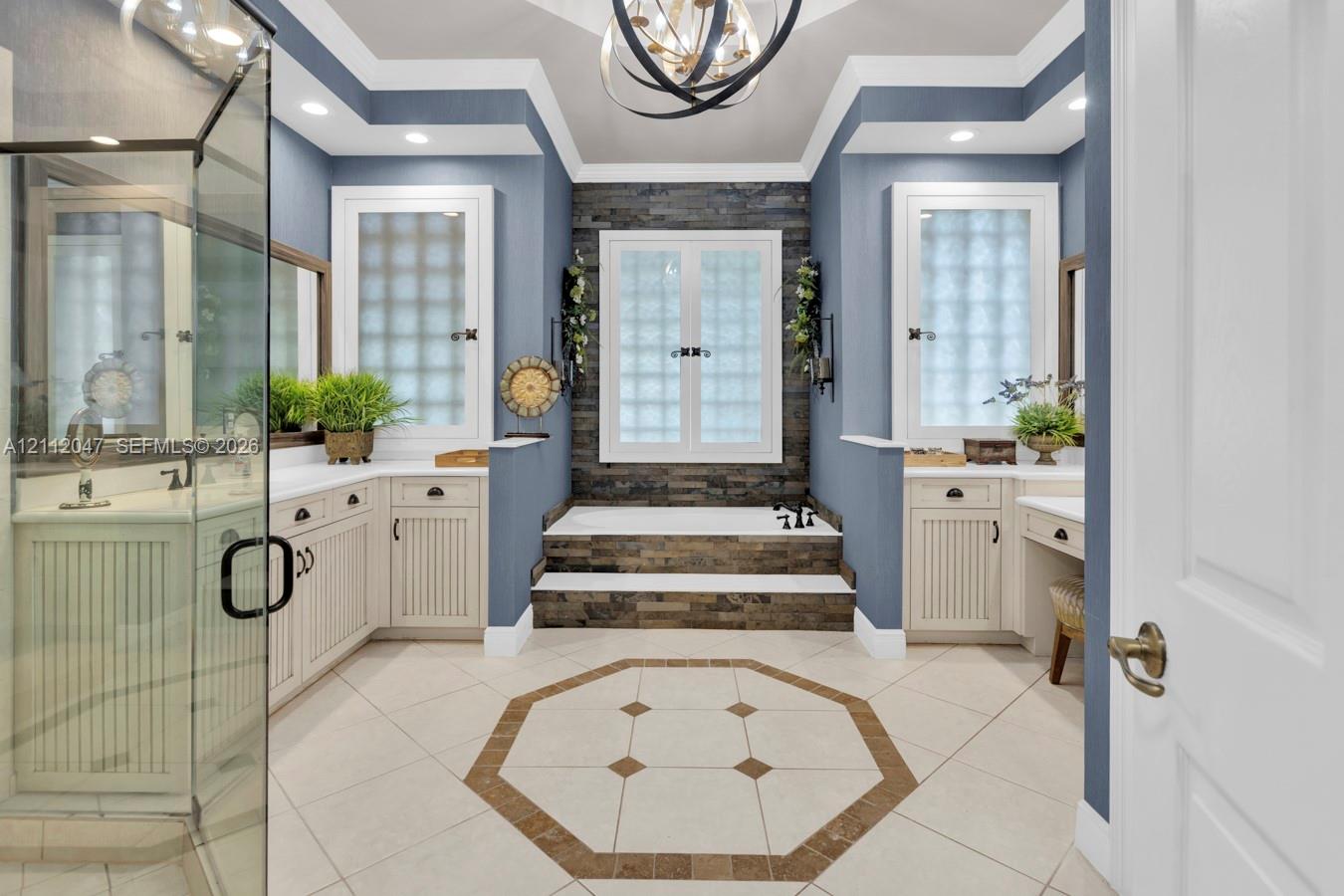 Bathroom featuring a raised soaking tub with stone-tile surround, dual white vanities with beadboard detailing, glass-enclosed shower, tray ceiling with statement chandelier, and geometric tile flooring with decorative inlay