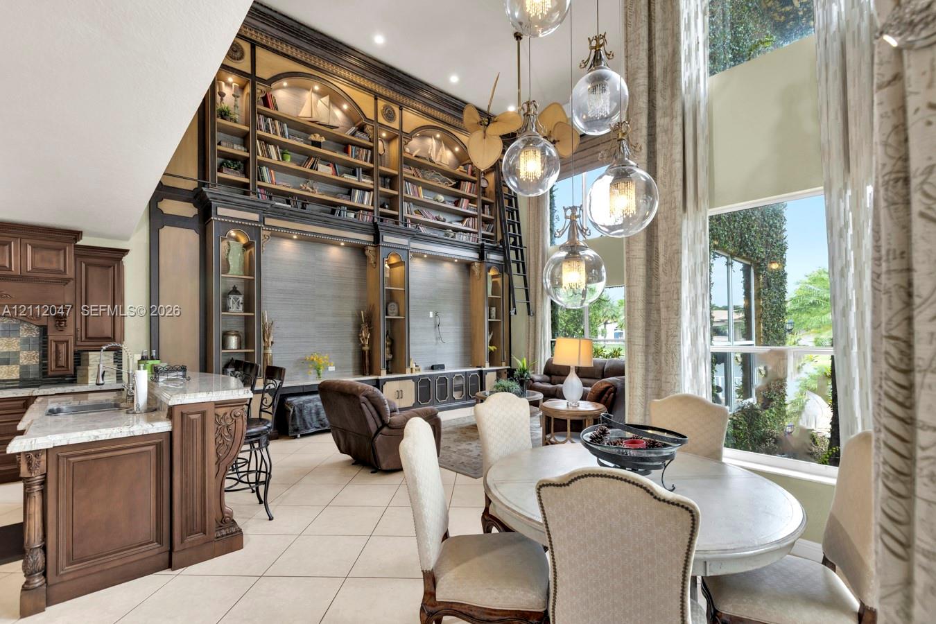 Two-story floor-to-ceiling built-in wooden shelving with integrated ladder, vaulted windows, multi-pendant orb lighting, and stone-surfaced island with ornate wood cabinetry