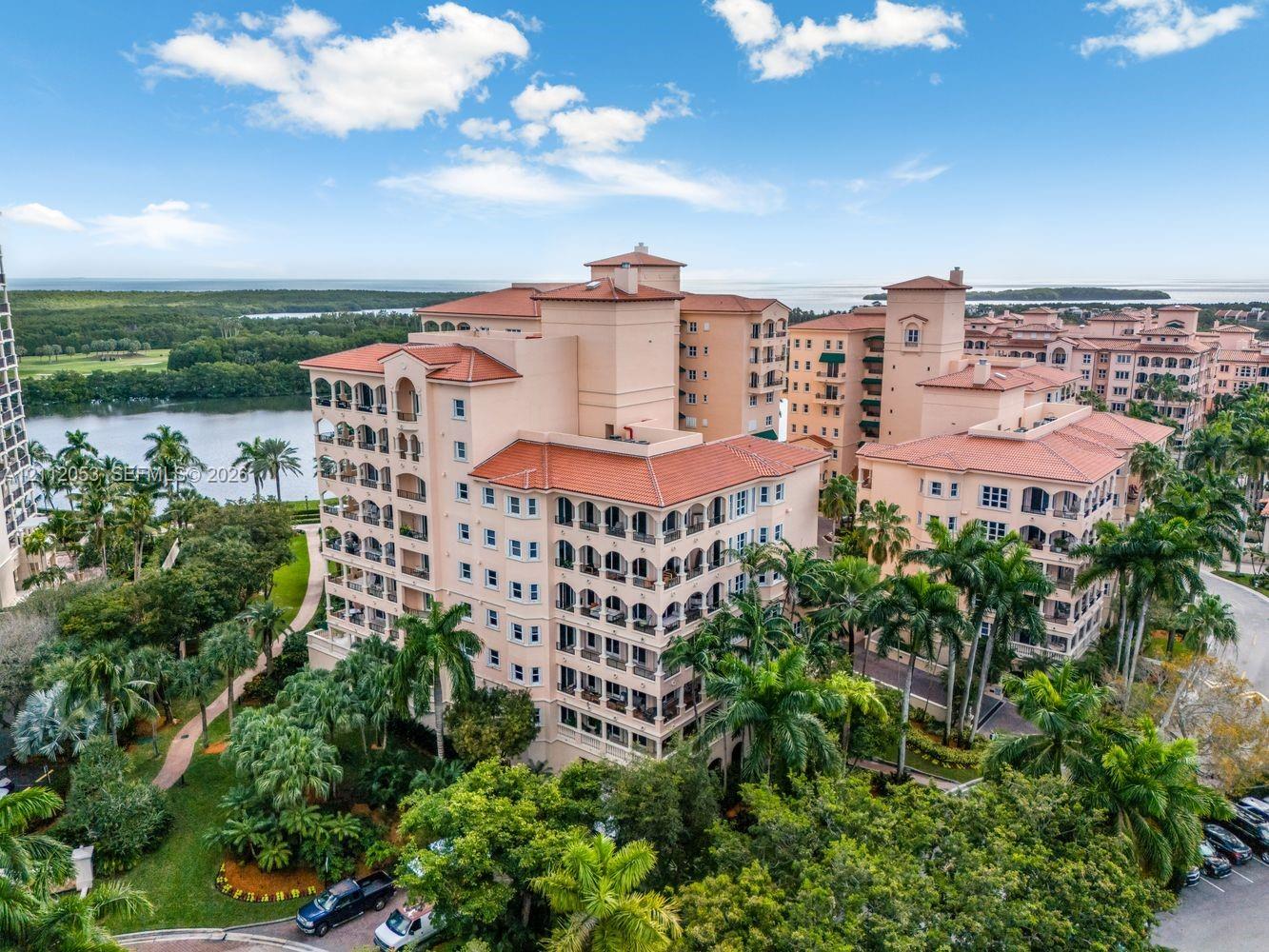 Multi-story Mediterranean-style complex featuring red tile roofing, recessed arched balconies, and exterior stucco finishes, situated amidst lush tropical landscaping with water frontage