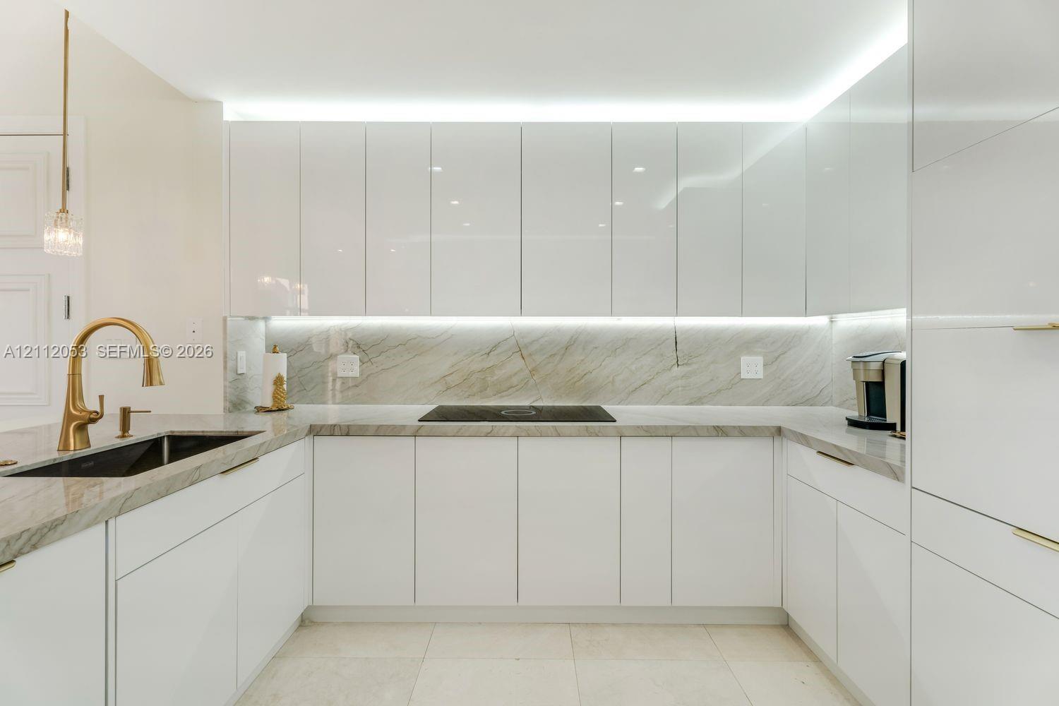 High-gloss white flat-panel cabinetry, stone countertops and backsplash with veined detailing, gold-tone faucet, under-cabinet lighting, and integrated cooktop