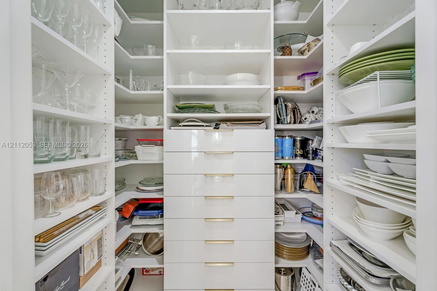 Custom walk-in pantry featuring white floor-to-ceiling shelving, tiered corner storage, and a central stack of drawers with metallic hardware