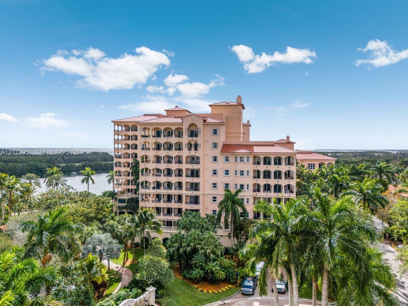 Mid-rise residential building featuring a terracotta tile roof, arched exterior balconies, lush tropical landscaping, and a waterfront setting