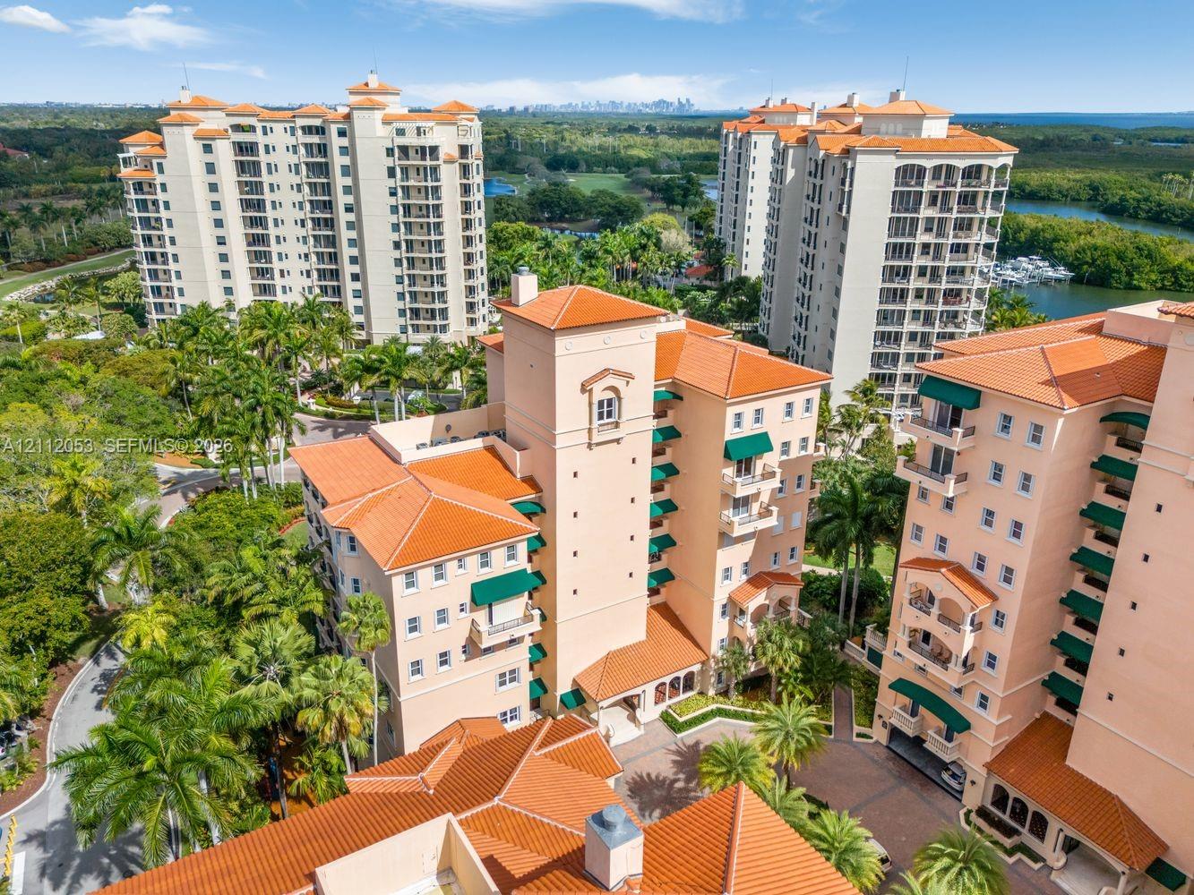Aerial perspective of mid-rise residential structures featuring terracotta-style roof tiles, light-toned stucco facades, integrated balconies with green awnings, and landscaped grounds with mature palm trees