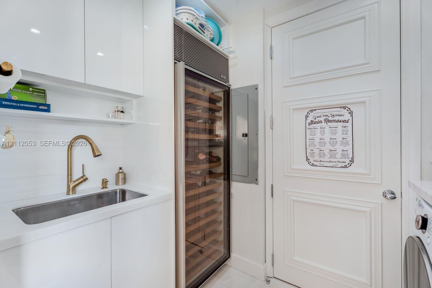 Built-in wine refrigerator, brass-tone gooseneck faucet, integrated stainless steel sink, custom white cabinetry, and multi-panel interior door