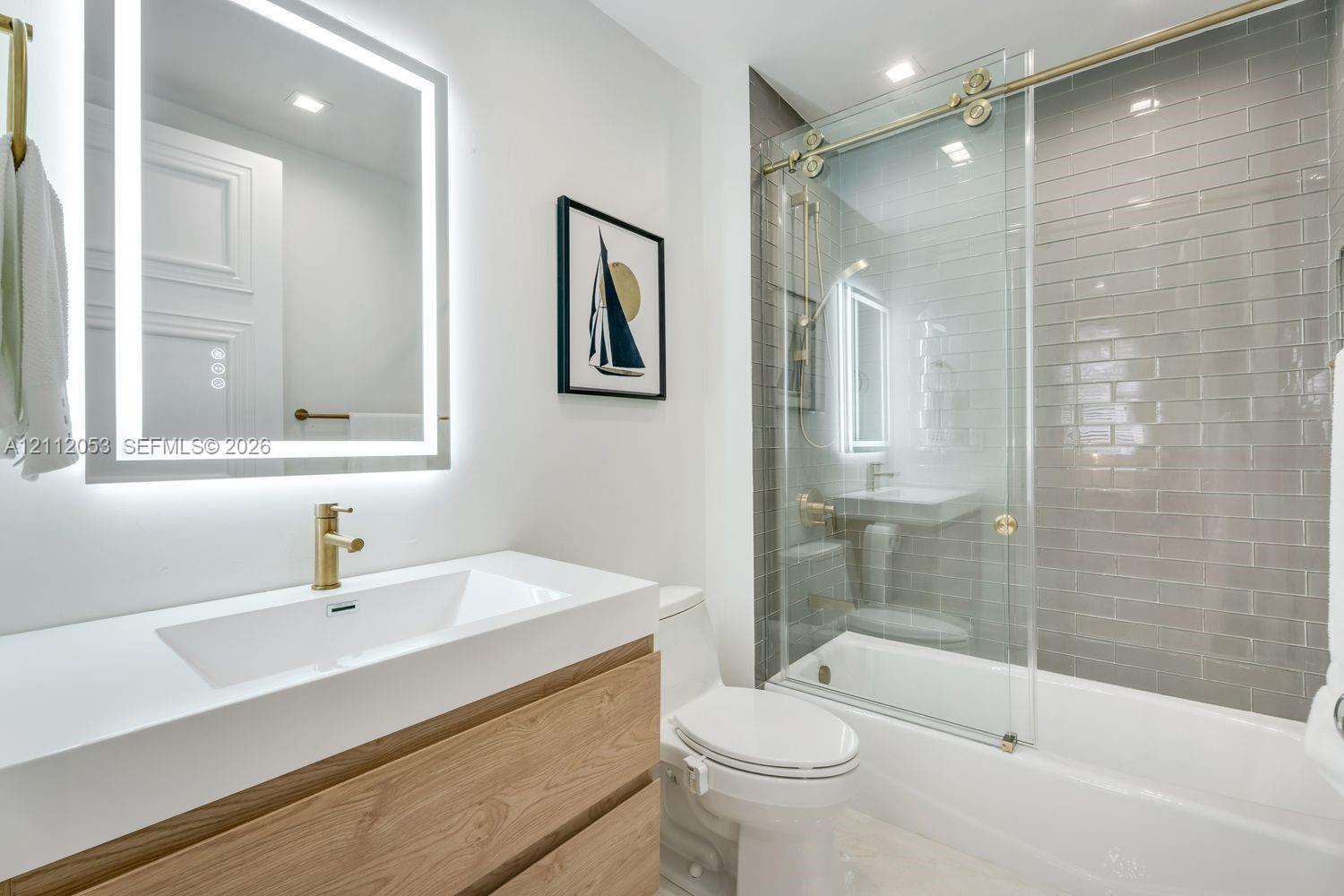 Floating wood-finish vanity with a solid surface countertop, brass faucet, LED-backlit mirror, and tub enclosure featuring sliding glass doors and ceramic wall tile