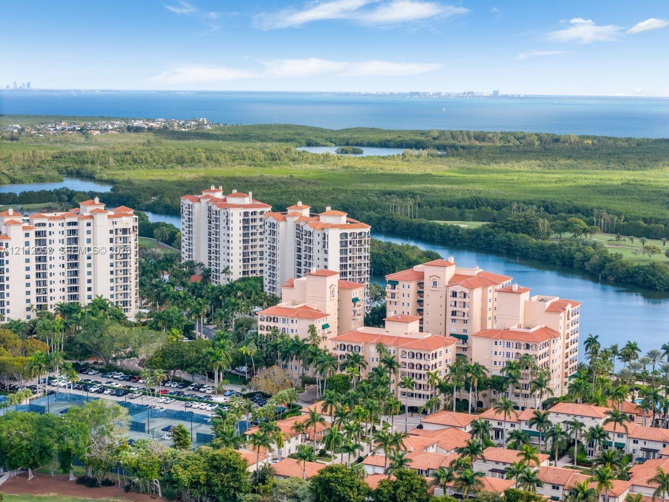 Aerial perspective of high-rise condominium complex featuring terracotta-tiled roofs, manicured palm landscaping, on-site tennis courts, and expansive water frontage