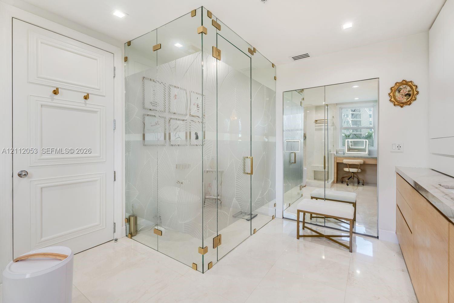 Frameless glass shower enclosure with gold-tone hardware, polished stone-finish flooring, custom light-wood cabinetry, and full-length mirrored panels