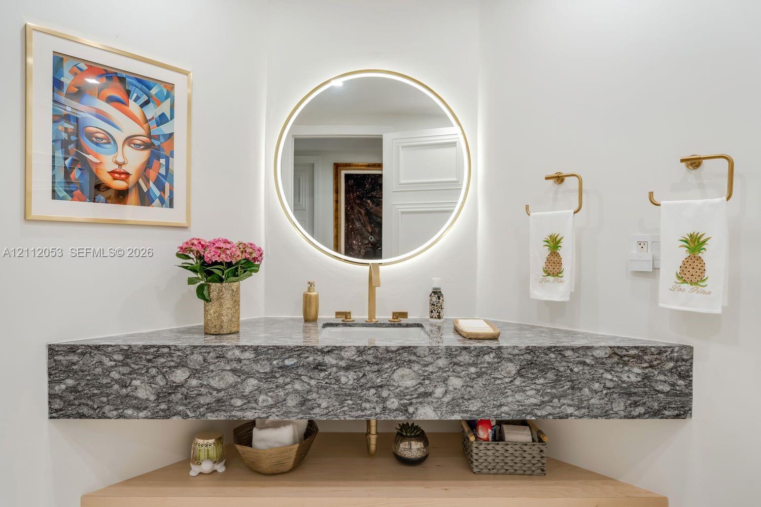 Corner-mounted floating vanity with stone countertop, integrated sink, brass-finish hardware, and round backlit mirror
