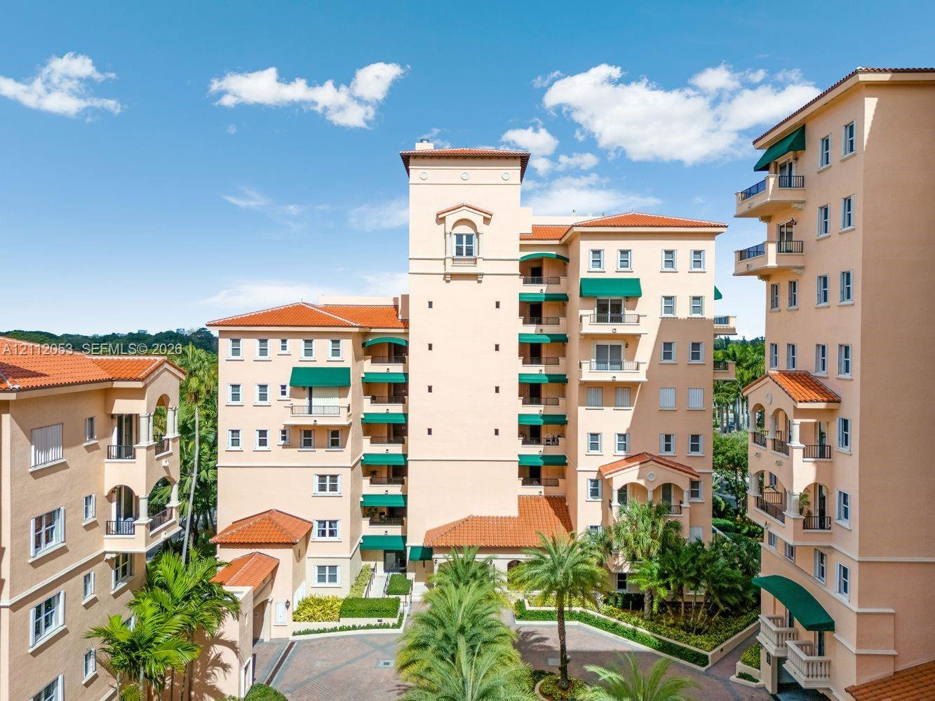Mediterranean-style multi-story buildings featuring terracotta tile roofs, stucco facades, integrated balconies with green fabric awnings, and a paved courtyard with mature palm trees