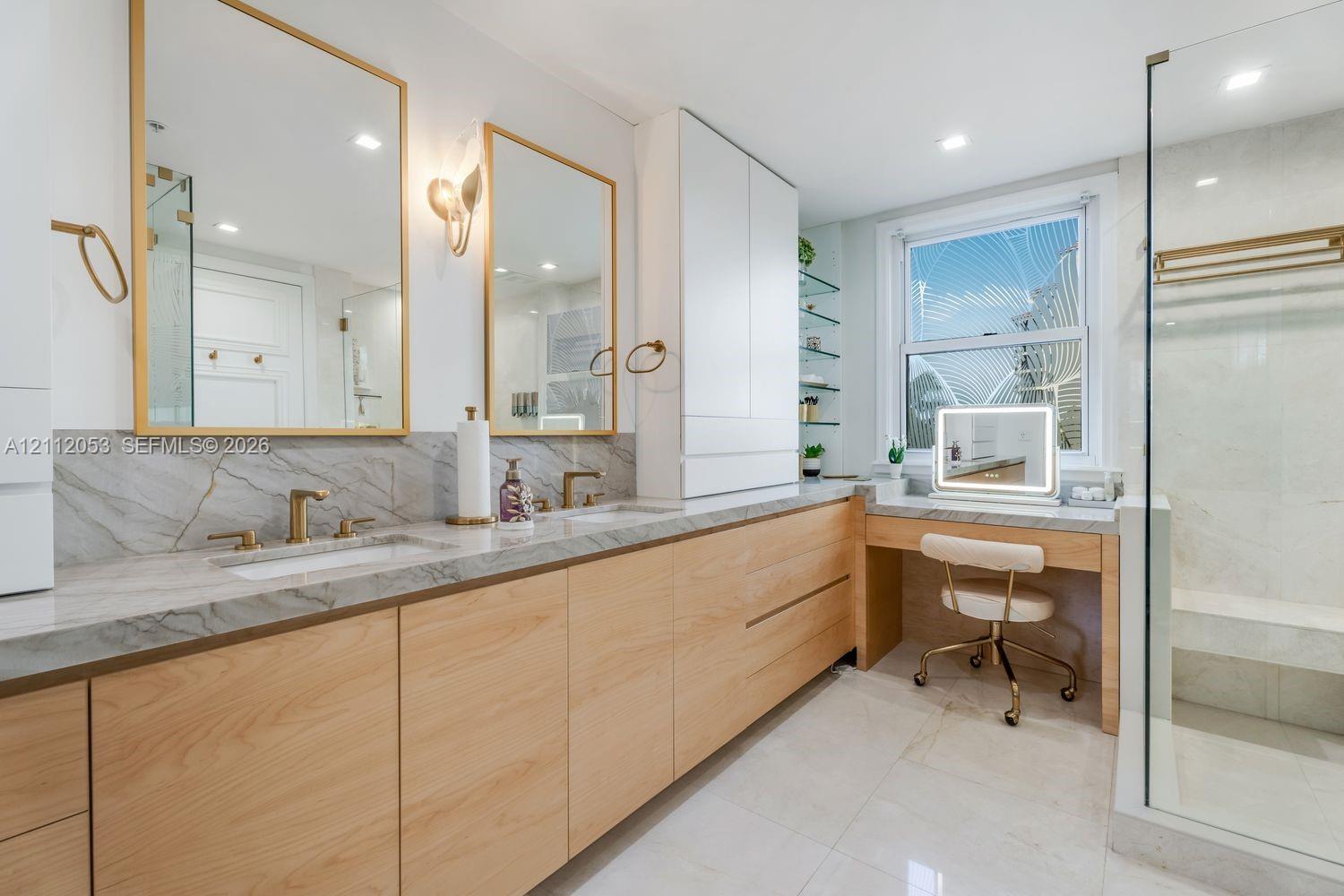 Dual-sink vanity with wood-finish cabinetry, stone countertop, gold-tone hardware, wall-mounted mirrors, and a glass-enclosed shower