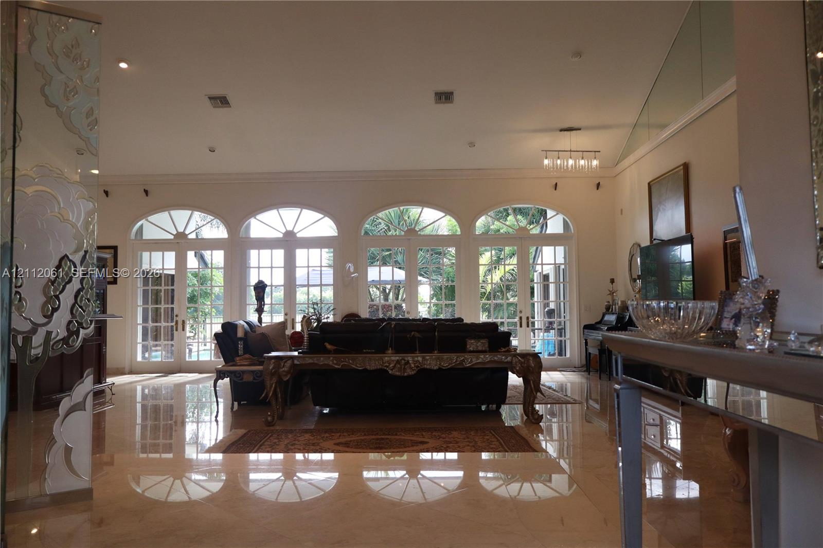 Open-concept living space featuring high ceilings, polished stone flooring, four arched French doors with fanlight transoms, and a glass-paneled staircase railing Open-concept living space featuring high ceilings, polished stone flooring, four arched French doors with fanlight transoms, and a glass-paneled staircase railing