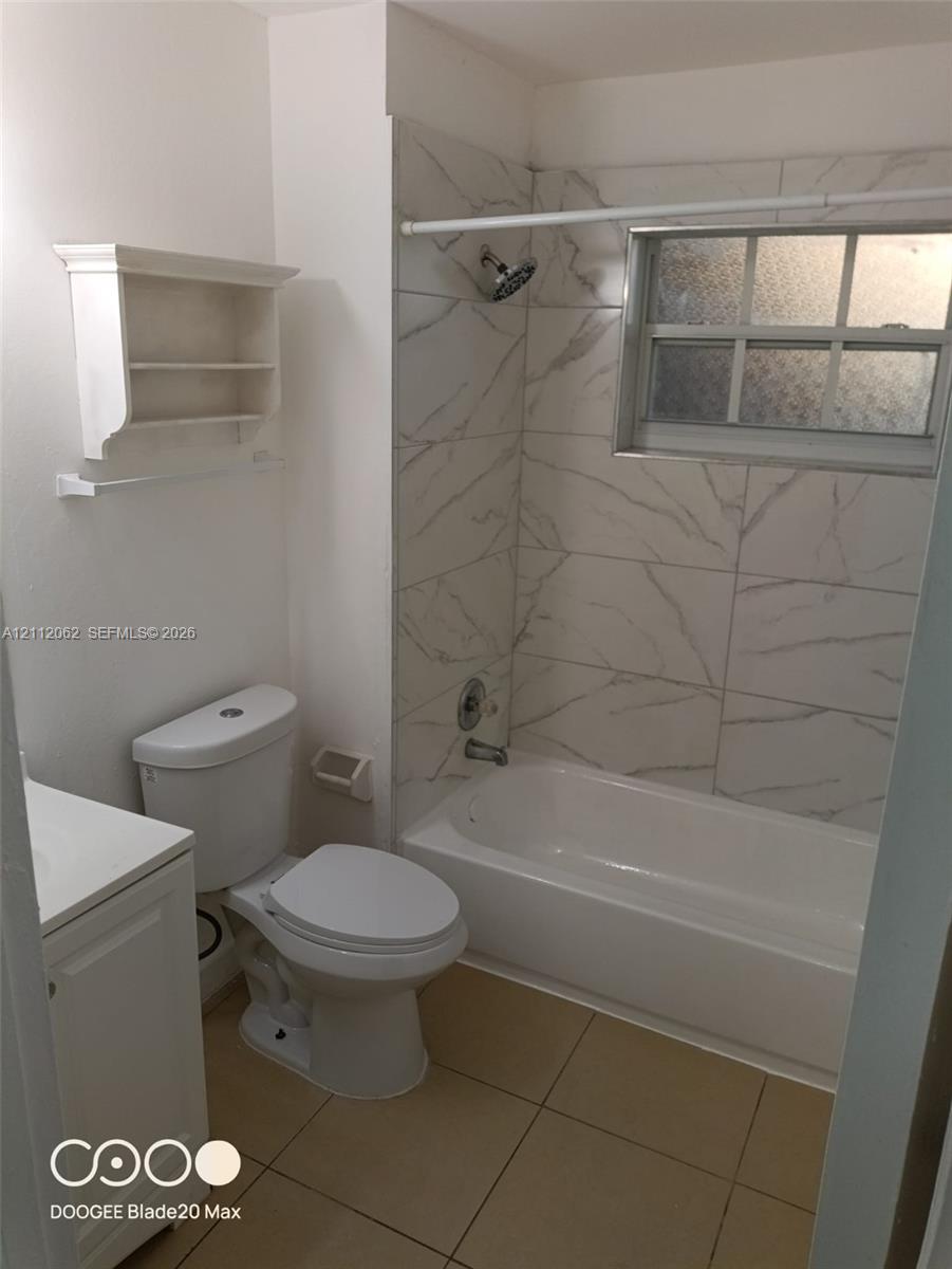 Full bathroom featuring a tub-shower combination with large-format, stone-patterned wall tiling, light-toned ceramic floor tile, a white vanity cabinet, and a built-in wall-mounted shelving unit
