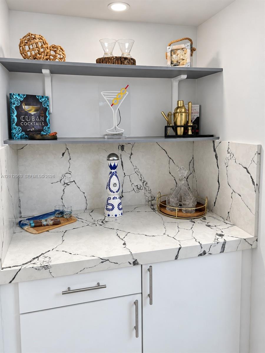 Built-in wet bar featuring stone-patterned surfacing, integrated backsplash, white cabinetry with metallic hardware, and dual grey floating shelves with recessed lighting Built-in wet bar featuring stone-patterned surfacing, integrated backsplash, white cabinetry with metallic hardware, and dual grey floating shelves with recessed lighting