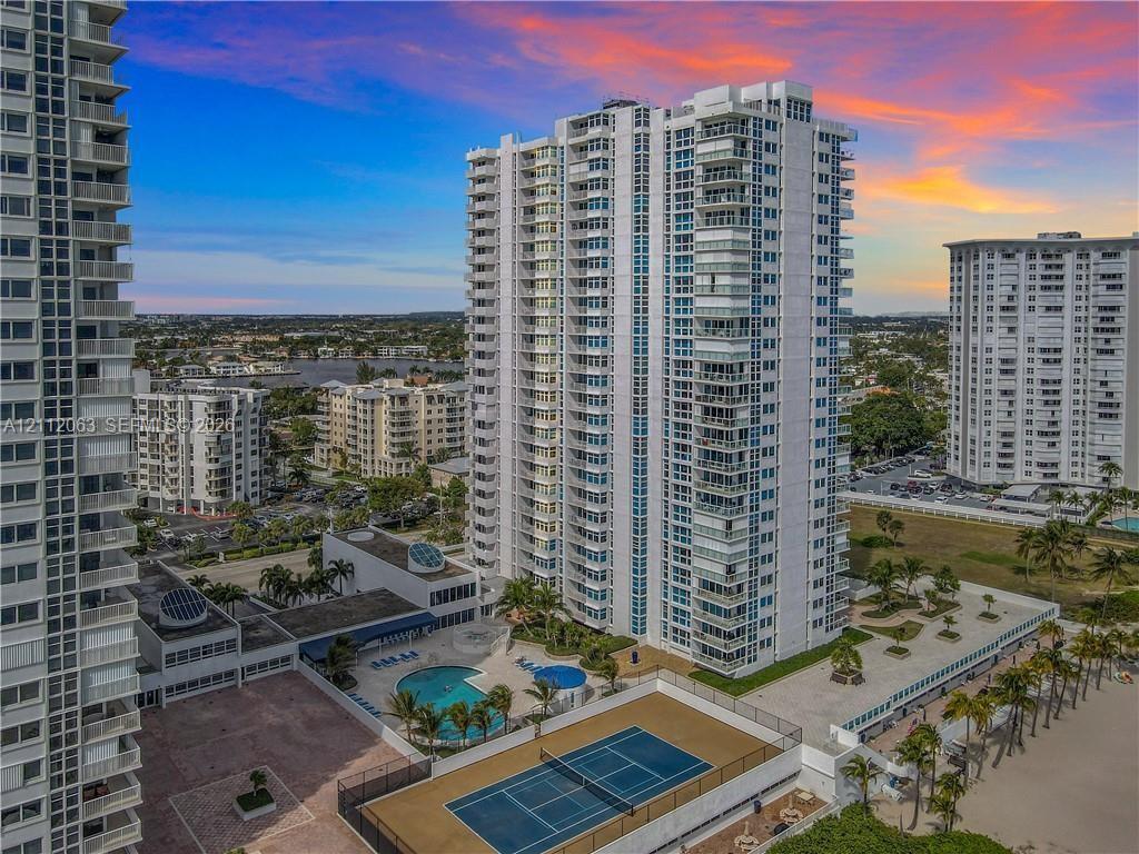 High-rise residential tower featuring private balconies, an outdoor swimming pool, a dedicated tennis court, and manicured tropical landscaping