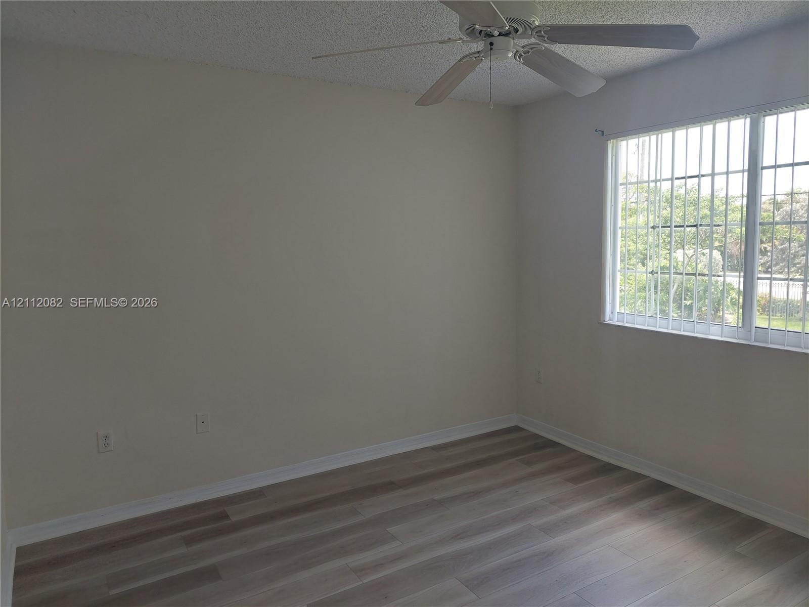 Room featuring wood-finish flooring, a multi-blade ceiling fan, and a window with security bars