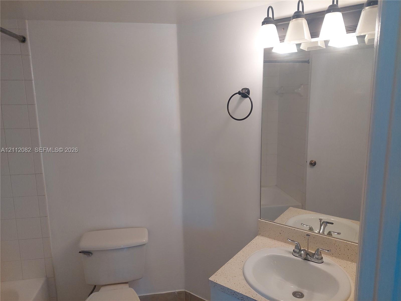 Bathroom featuring a vanity with a solid-surface countertop, an integrated oval sink, a wall-mounted light fixture with four frosted glass shades, a large plate glass mirror, and a wall-mounted towel ring