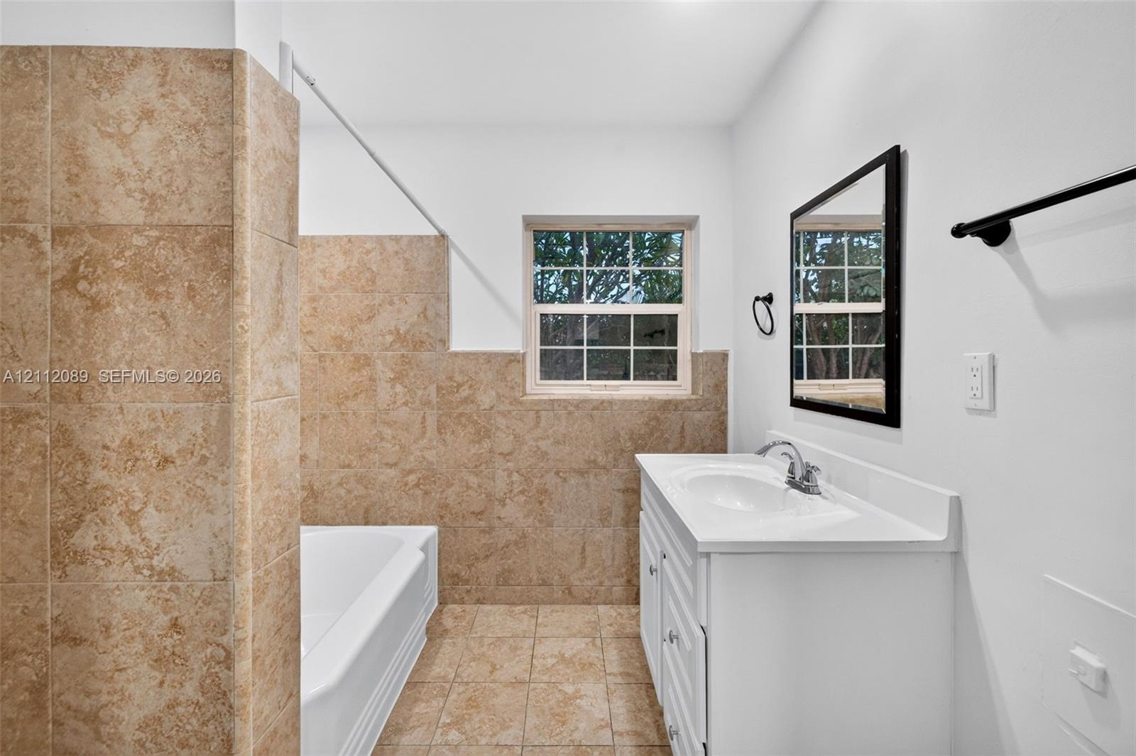 Bathroom featuring a white vanity with an integrated sink, a deep soaking tub with a tiled surround, neutral floor tiling, a framed wall mirror, and matte black fixtures