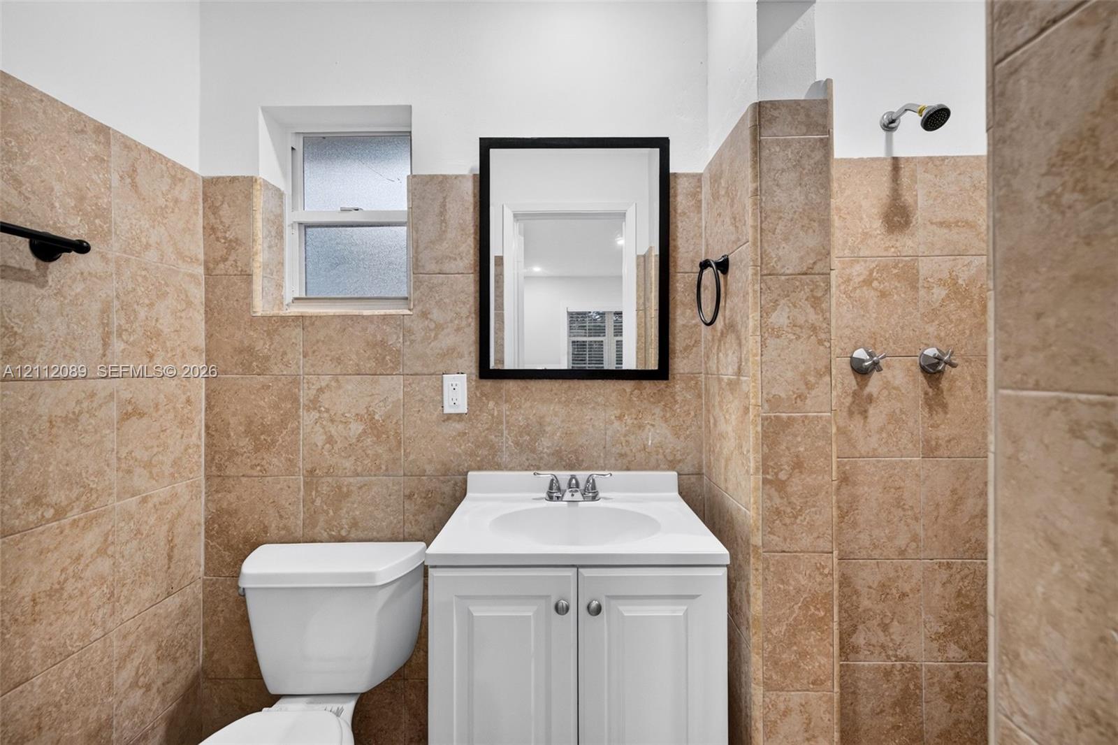 Bathroom featuring tan stone-tile wall cladding, a white pedestal vanity with an integrated sink, a framed wall mirror, and a wall-mounted shower head