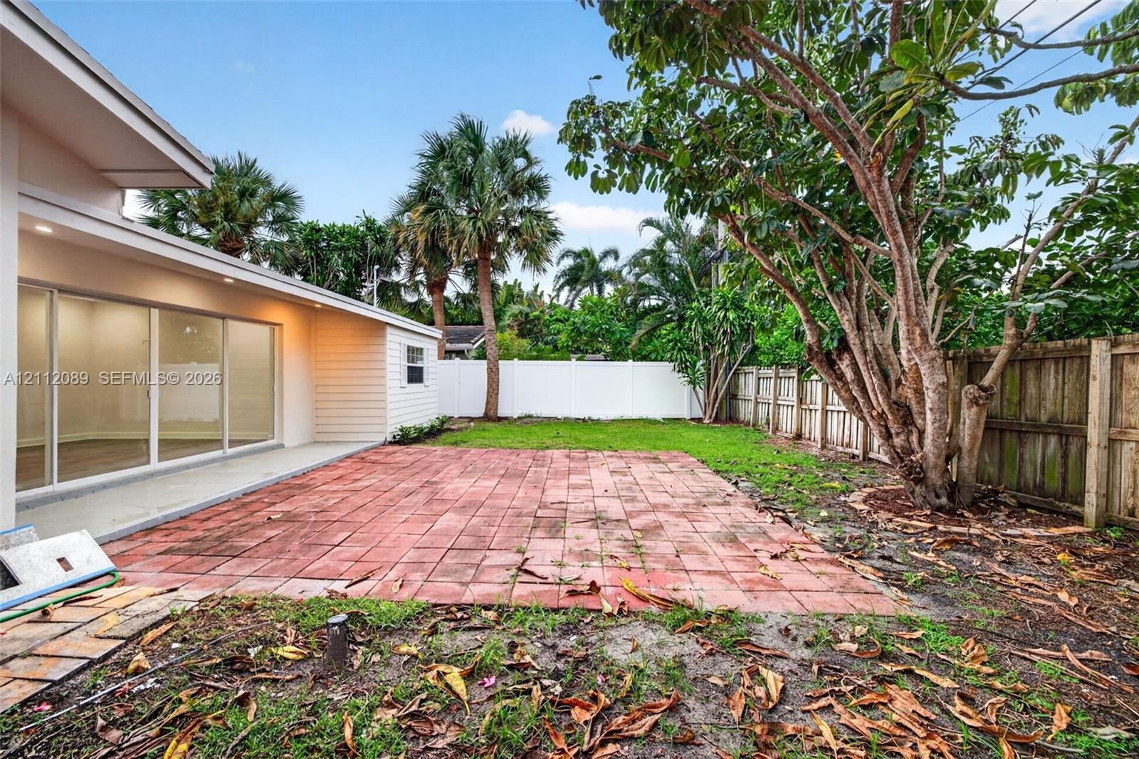 Backyard featuring a red brick patio, white vinyl privacy fencing, mature trees, and glass sliding door access