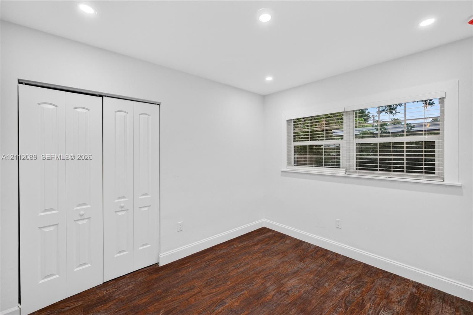 Interior room featuring dark wood-finish flooring, recessed ceiling lighting, a large window with horizontal blinds, and a double-panel closet with raised-panel doors