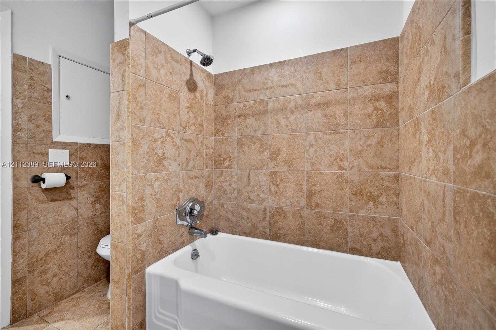 Full-height tan stone-finish tile surround, white acrylic soaking tub, wall-mounted showerhead, and integrated chrome faucet hardware