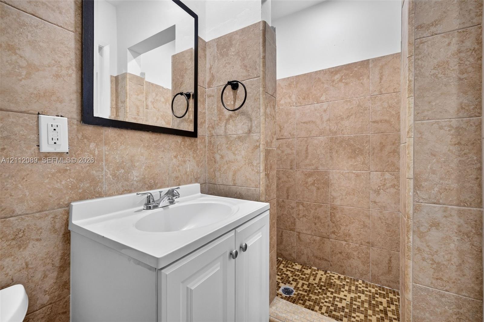 White vanity with integrated basin, chrome-finish dual-handle faucet, wall-mounted black-framed mirror, and full-height stone-finish tile surround with a mosaic tile shower floor