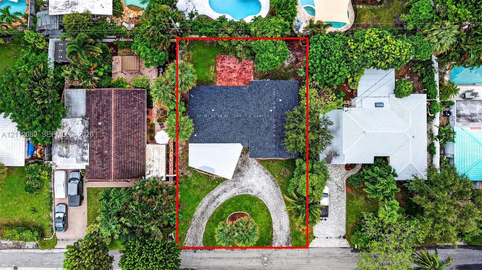 Aerial perspective of property featuring a dark-toned shingle roof, a circular driveway, mature perimeter landscaping, and a rear stone-paved patio